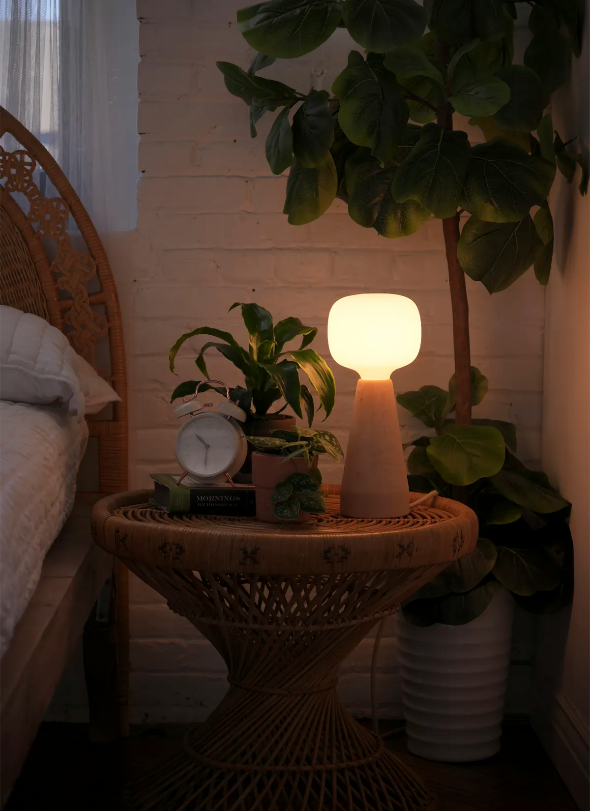 An Aura™ Ambient Grow Light with a natural wood base sits atop a bedside table softly lighting a two small potted plants. 