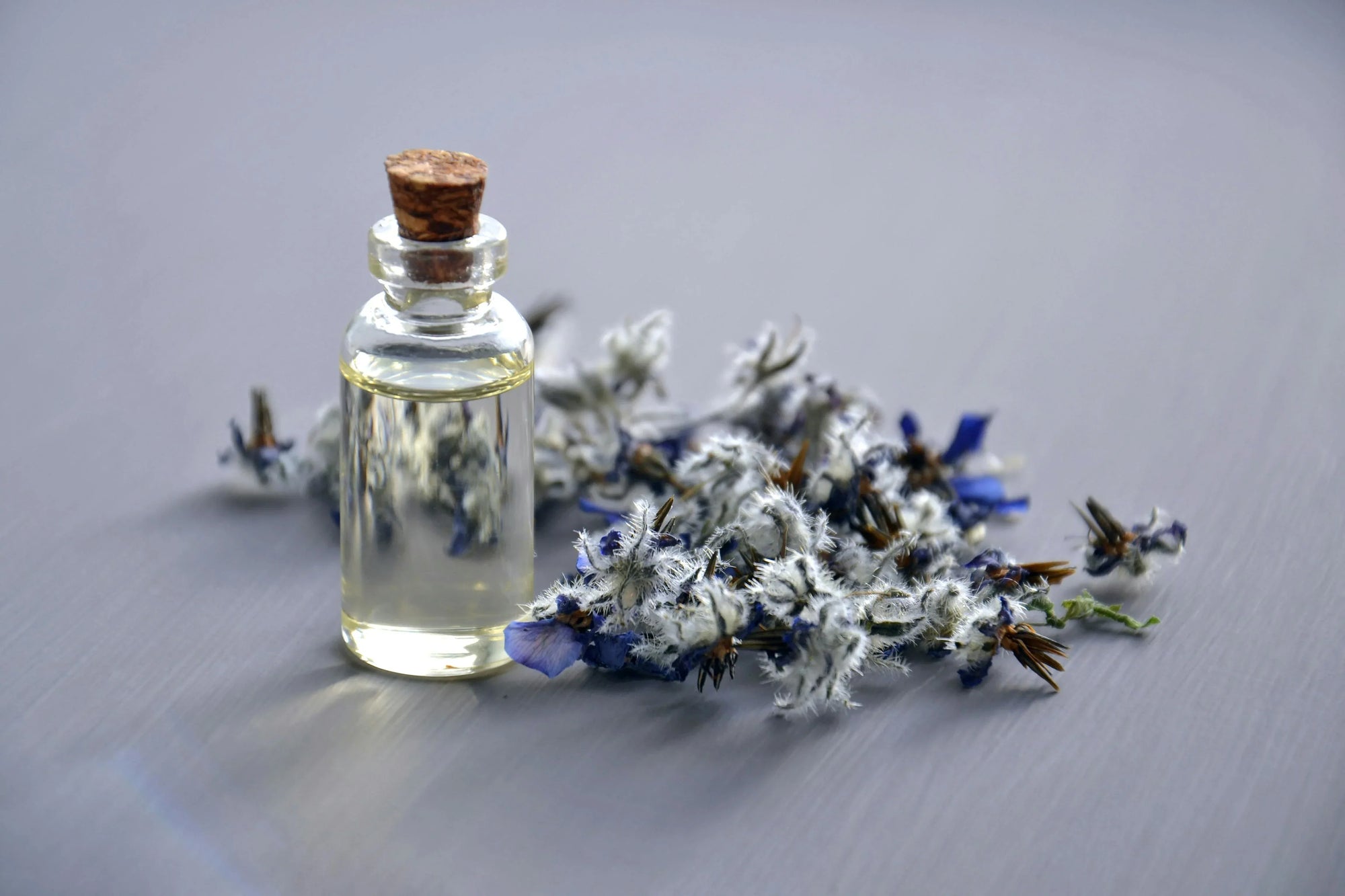 A small glass bottle with cork, filled with clear liquid, sits on a gray surface. Beside it, a cluster of delicate blue and white flowers evokes tranquility.