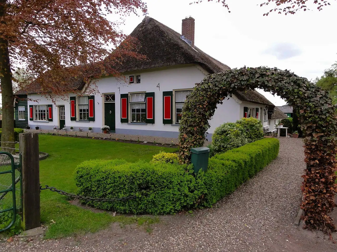A charming house with red shutters and a thatched roof is surrounded by lush greenery. A leafy archway leads to a gravel path, creating a cozy, inviting atmosphere.