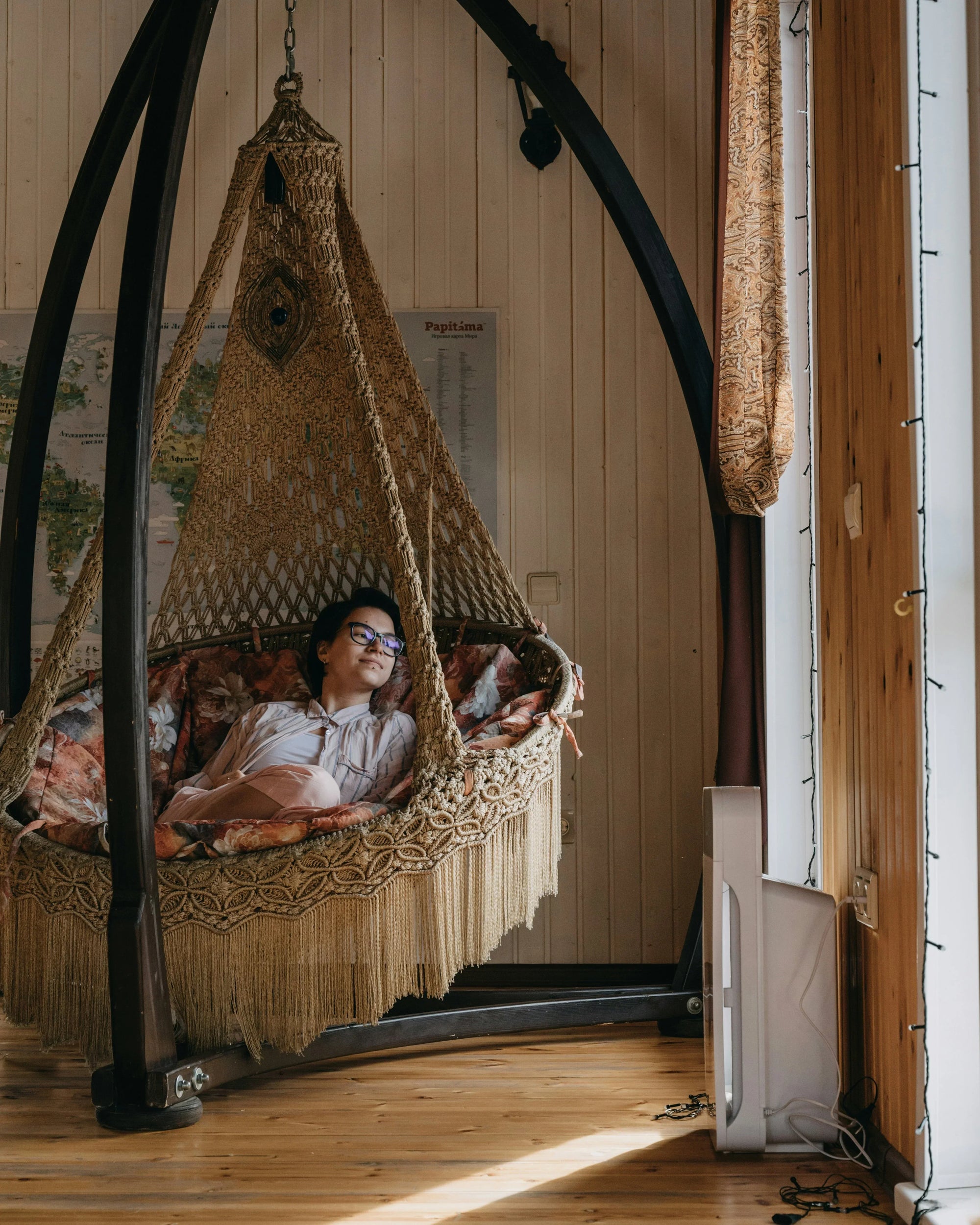A person wearing sunglasses relaxes in a cozy, woven hammock chair inside a warmly lit room. The setting feels tranquil and inviting.