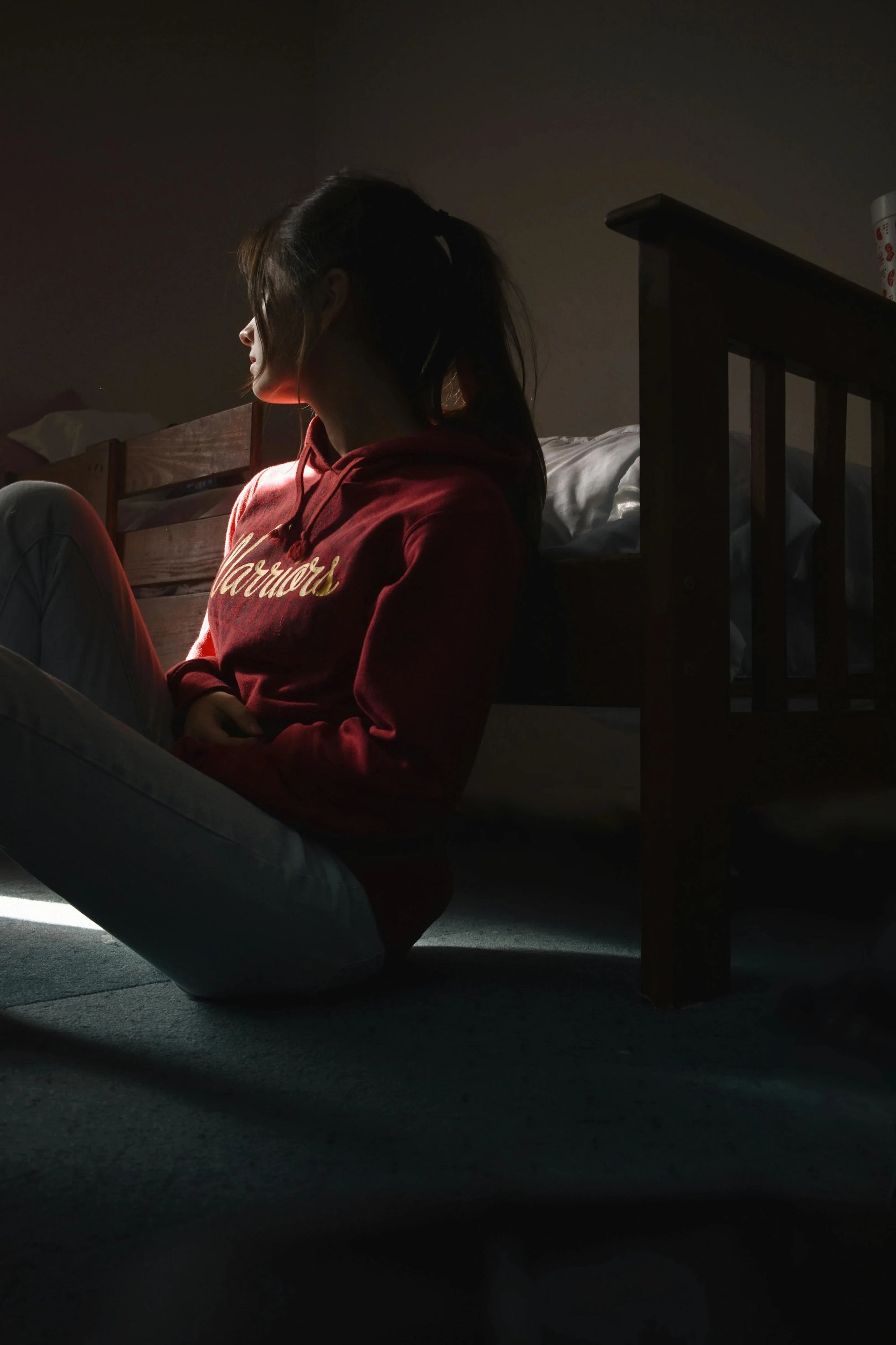 A young woman in a red "Warriors" hoodie sits on the floor in a dimly lit room, facing left. Sunlight from a window casts shadows, creating a contemplative mood.