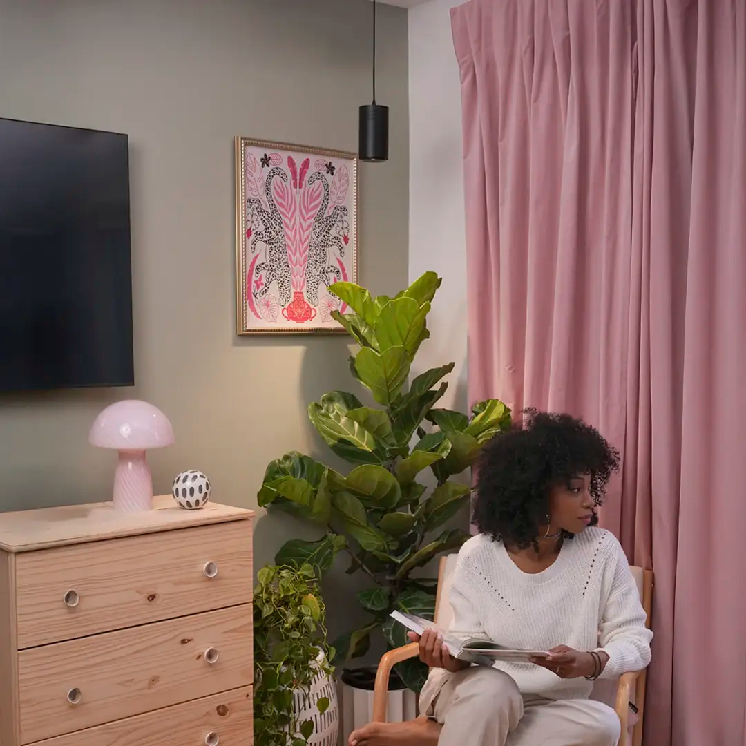 A woman in a cozy room sits on a wooden chair, holding a magazine. The setting features pink curtains, a potted plant, a light wood dresser with a pink lamp, and a colorful framed artwork. The atmosphere is calm and inviting.