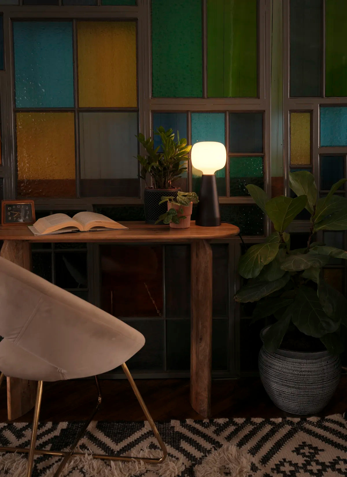 Dimly lit room illuminated with a Soltech Aura on a desk with a book, clock and potted plants. The background is stained glass.
