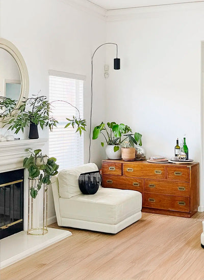 A cozy living room corner featuring a white armchair with a black vase, wooden dresser with green plants, a round mirror, and wine bottles and a wall mounted Soltech Aspect exuding a serene, elegant ambiance.