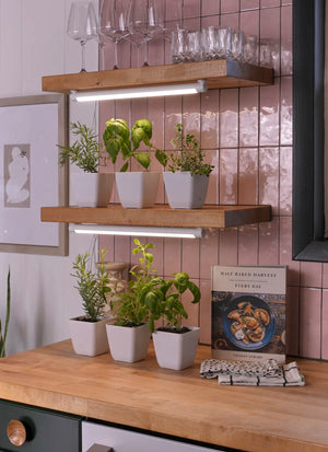 Wooden shelves on a pink tiled wall hold glasses and potted herbs. Soltech Groves mounted above each shelf. A cookbook and kitchen towel rest on a wooden counter below, creating a cozy kitchen vibe.