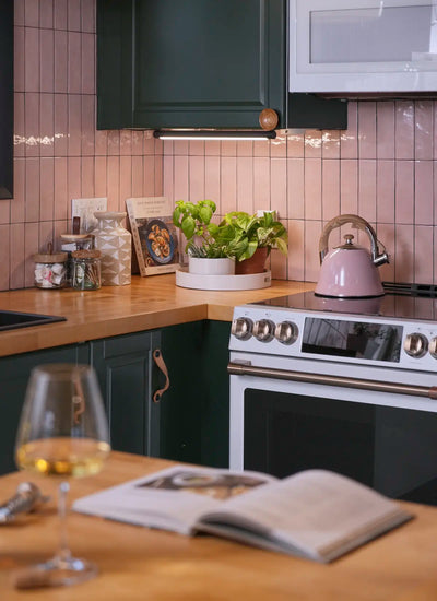 Cozy kitchen with green cabinets, pink tiles, and wooden countertops. A pink kettle sits on the stove; fresh herbs are sitting underneath a mounted Soltech Grove and a cookbook is nearby. A glass of white wine and an open book create a relaxed vibe.