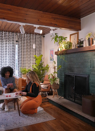 Modern living room with two women playing cards at a wooden coffee table. The setting has a cozy feel, with plants, a black brick fireplace, dotted curtains and Soltech Highland tracking system mounted above them.
