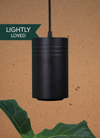 A black large Soltech Aspect growlight pendant hanging against a brown background with green foliage to the side.