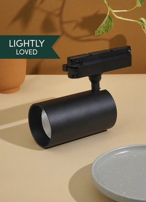 Black Soltech Highland light fixture on a beige surface with a plant and pot in the background. Label that says Lightly Loved"