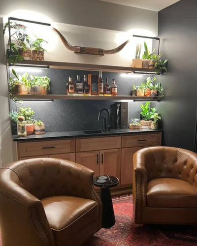 Modern kitchen with leather chairs, shelves with plants illuminated by Soltech Versas and bottles, and a dark backsplash.
