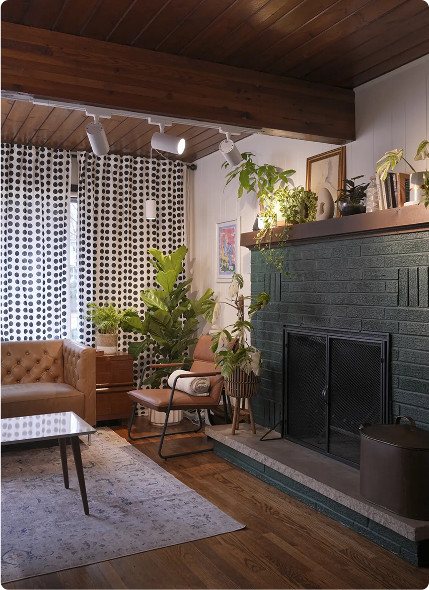 A cozy living room with a mid-century modern vibe, featuring a brown leather sofa, green brick fireplace, various plants illuminated by the Soltech Highland Track Light System, and polka dot curtains.