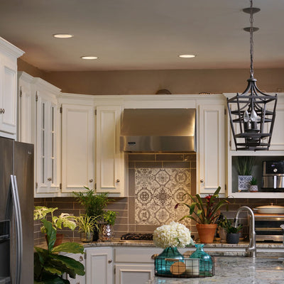 A cozy kitchen with white cabinets and a decorative tile backsplash. Features include a metal range hood, potted plants, and pendant lighting.
